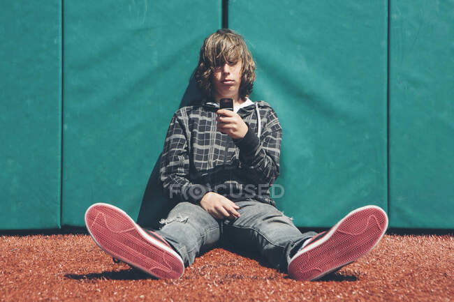 Adolescente sentado contra la pared acolchada en el campo de deportes, sosteniendo el teléfono móvil - foto de stock