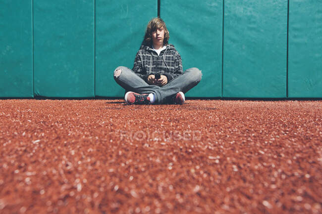 Ragazzo adolescente seduto contro il muro imbottito al campo sportivo, tenendo il telefono cellulare — Foto stock
