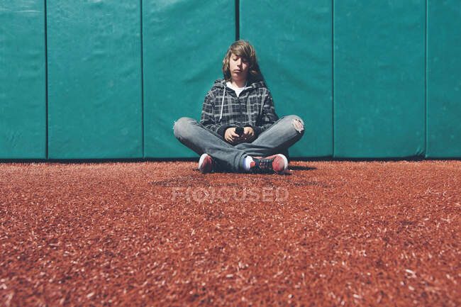 Adolescente sentado contra a parede acolchoada no campo de esportes, segurando telefone celular — Fotografia de Stock