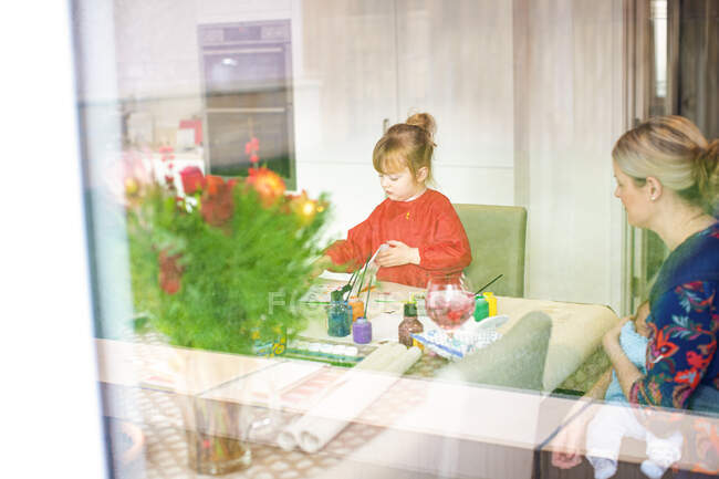 Young girl using paints at kitchen table with mother sitting nearby holding baby — Stock Photo