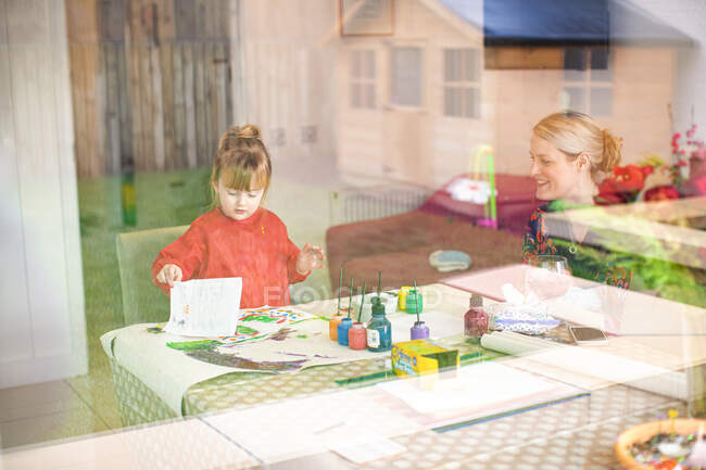 Молода дівчина використовує фарби за кухонним столом з матір'ю, яка сидить поруч тримає дитину — стокове фото