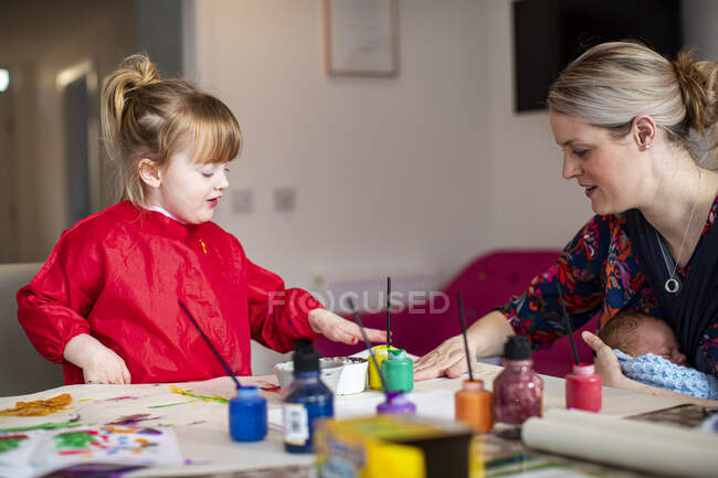 Young girl using paints at kitchen table with mother sitting nearby holding baby — Stock Photo