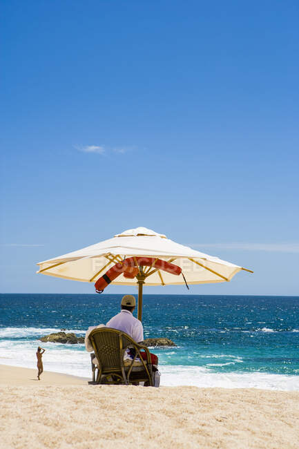Вид сзади человека, сидящего под зонтиком на белом песчаном пляже у океана — стоковое фото