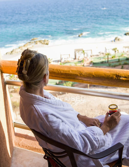 Adult woman sitting with a drink on a hotel balcony overlooking a blue ocean and white sand beach — Stock Photo