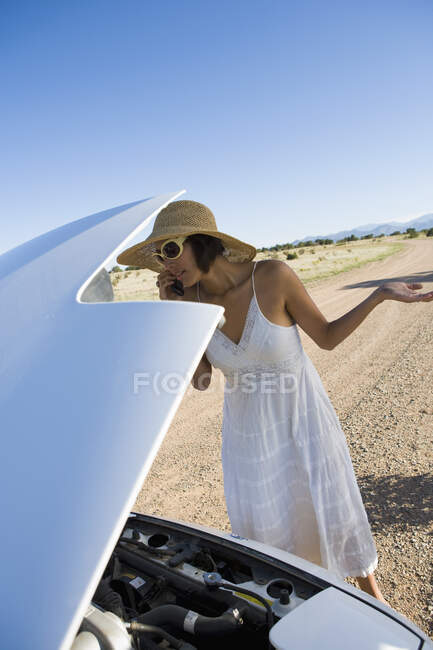 Mujer nativa americana en vestido de sol conduciendo un coche deportivo convertible blanco en el desierto camino de tierra que tiene problemas de coche - foto de stock