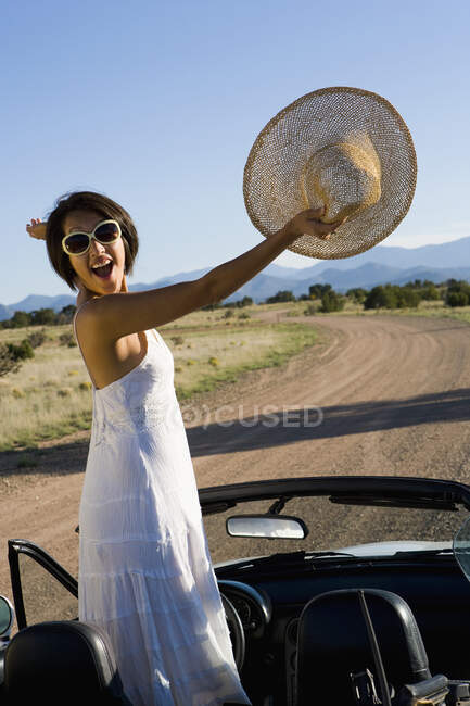 Mujer nativa americana en vestido de sol conduciendo un coche deportivo convertible blanco en el camino de tierra del desierto - foto de stock