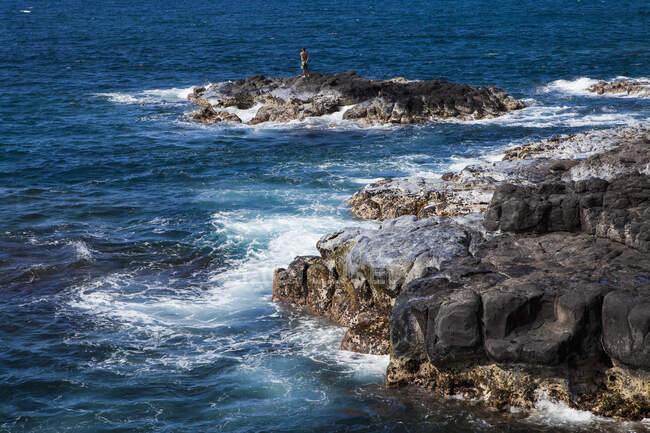 Rocas lavales negras en una costa dentada con olas rompiéndose, una persona de pie en un afloramiento. - foto de stock