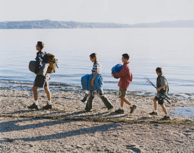 Famille de quatre personnes portant du matériel de camping, marchant sur la plage au crépuscule — Photo de stock