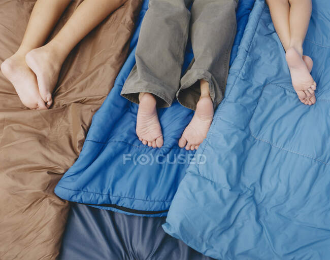 Tres personas, un adulto y dos niños acostados en sacos de dormir, rodillas abajo, pies desnudos. - foto de stock