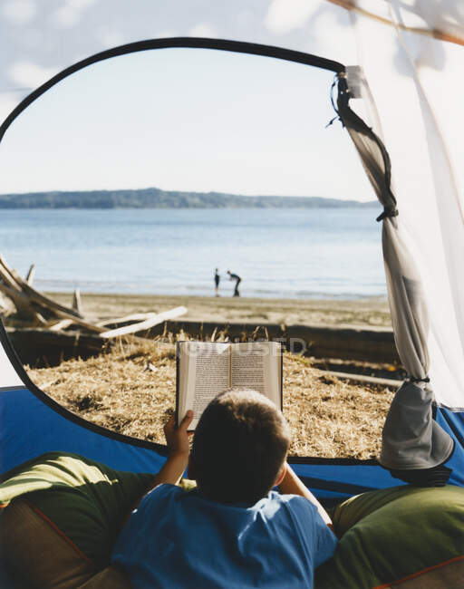 Young boy reading book at the opening of a tent on the beach. — Stock Photo