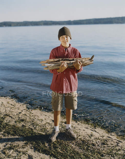 Portrait of adolescent boy holding pile of firewood on beach — Stock Photo