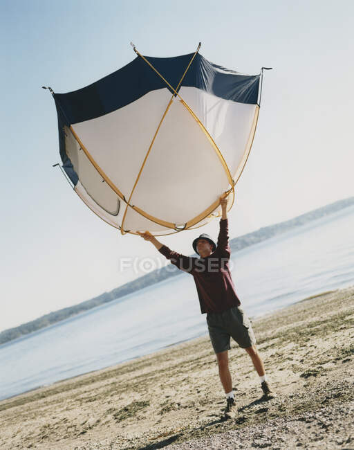 Hombre sosteniendo una pequeña tienda de cúpula sobre su cabeza, de pie en la playa - foto de stock