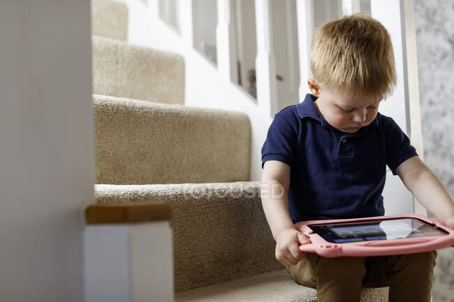 Three year old boy sitting on the stairs looking at a digital tablet. — Stock Photo