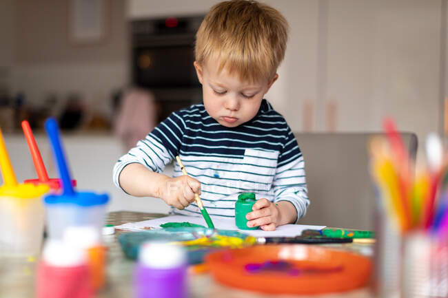 Garçon de trois ans occupé à peindre à la maison, avec des pots de peinture et des pinceaux. — Photo de stock