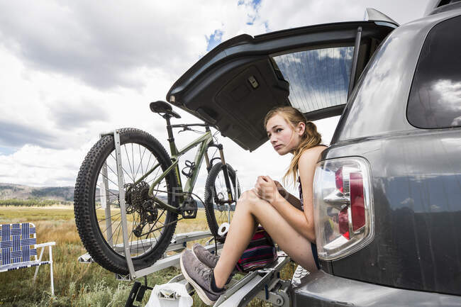 Adolescente sentada en la puerta trasera de un SUV mirando la vista - foto de stock