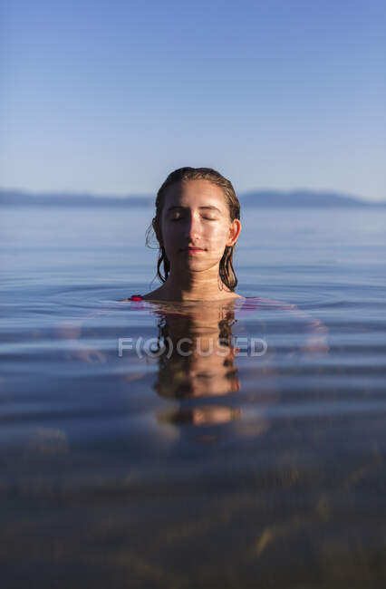 Adolescente con los ojos cerrados, cabeza y hombros por encima de las aguas tranquilas de un lago al amanecer - foto de stock