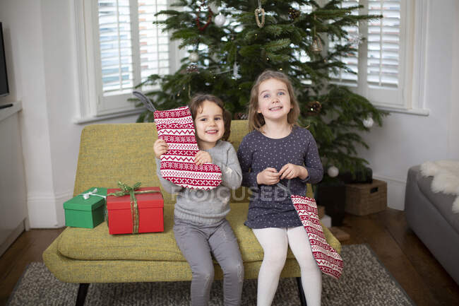 Две молодые девушки сидят на диване в гостиной, держа красный и белый рождественский чулок, улыбаясь в камеру. — стоковое фото