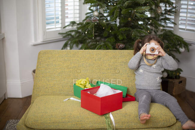 Молодая девушка сидит на диване в гостиной с рождественским подарком в красной коробке, фотографируясь с игрушечной камерой. — стоковое фото