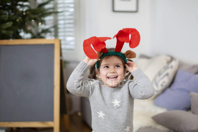 Smiling young girl wearing grey jumper putting on reindeer antler headband. — Stock Photo