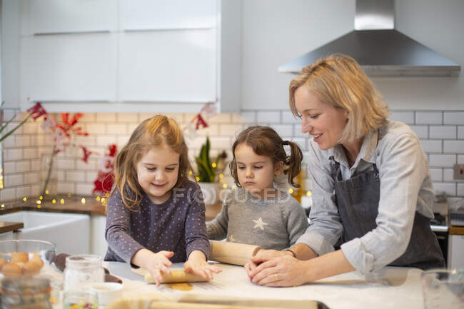 Blonde Frau mit blauer Schürze und zwei Mädchen, die in der Küche stehen und Weihnachtsplätzchen backen. — Stockfoto