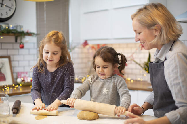 Donna bionda che indossa grembiule blu e due ragazze in piedi in cucina, cuocere i biscotti di Natale. — Foto stock