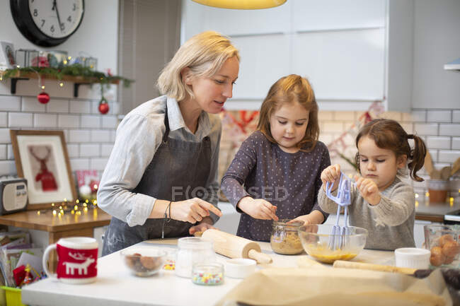 Mujer rubia con delantal azul y dos chicas de pie en la cocina, horneando galletas de Navidad. - foto de stock