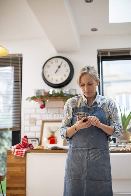 Блондинка в синьому фартусі стоїть на кухні, використовуючи мобільний телефон . — стокове фото