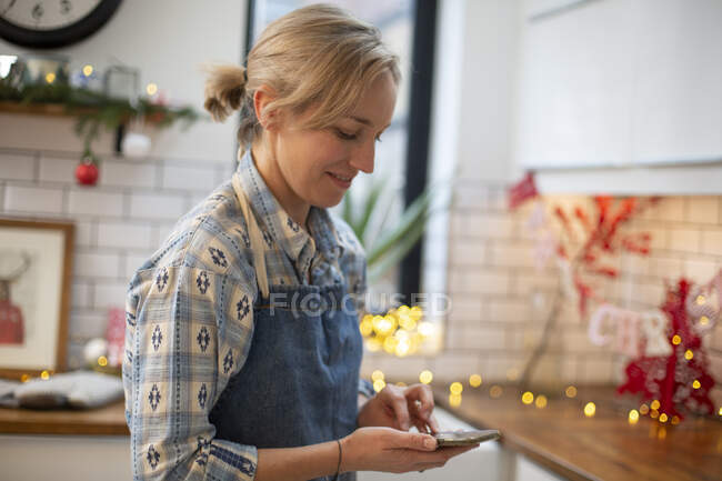 Mujer rubia con delantal azul de pie en la cocina, usando el teléfono móvil. - foto de stock