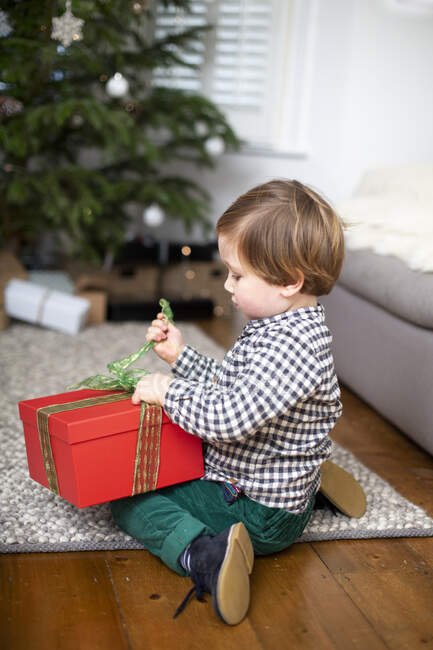 Niño sentado en el suelo de la sala de estar, desenvolviendo regalo de Navidad en caja roja. - foto de stock