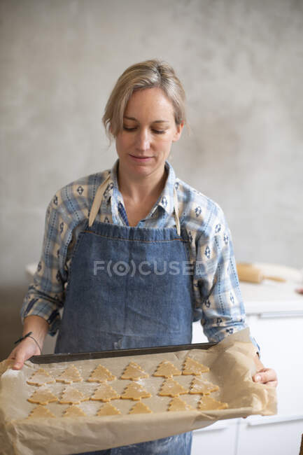Blond woman wearing blue apron holding tray with Christmas Tree cookies. — Stock Photo