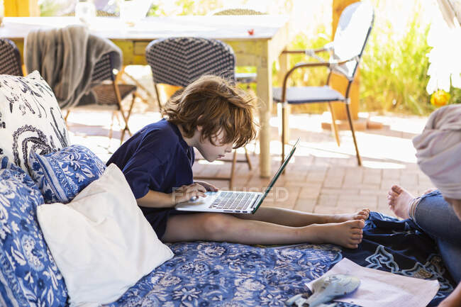 Ragazzo con i capelli castani seduto sul letto all'aperto, facendo i compiti sul computer portatile. — Foto stock