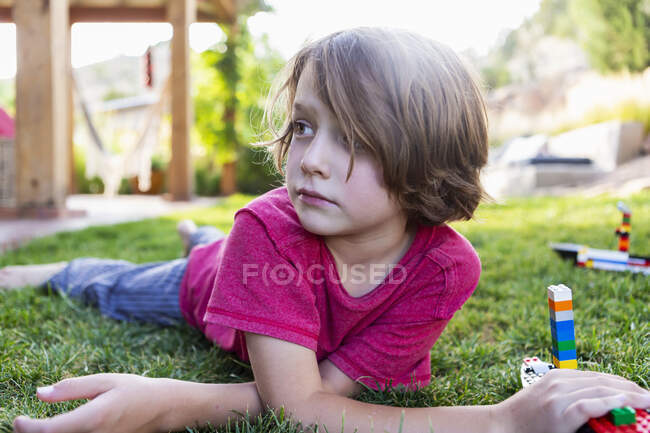 Хлопчик з коричневим волоссям лежить на газоні, грає з будівельними блоками . — стокове фото