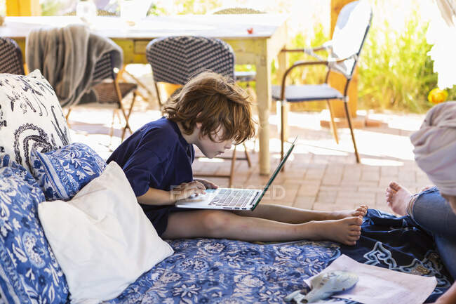 Niño con el pelo castaño sentado en la cama al aire libre, haciendo la tarea en el ordenador portátil. - foto de stock