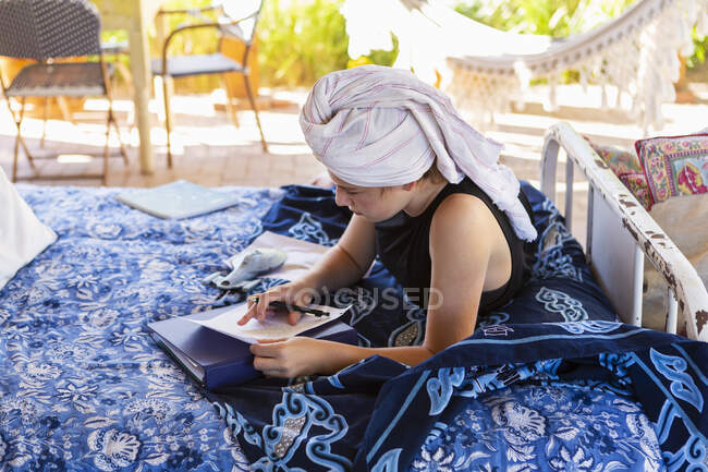 Adolescente con el pelo envuelto en toalla sentada en la cama al aire libre, haciendo la tarea. - foto de stock