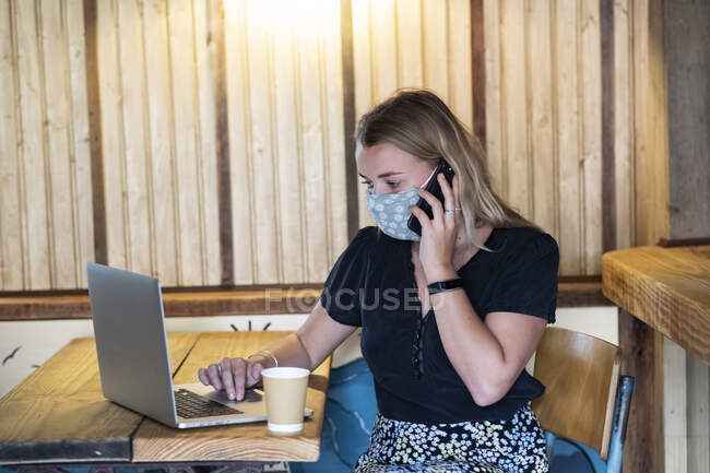 Jovem loira vestindo máscara facial azul, sentada à mesa, usando telefone celular e computador portátil. — Fotografia de Stock