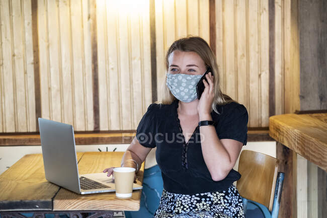 Jeune femme blonde portant un masque bleu, assise à table, utilisant un téléphone portable et un ordinateur portable. — Photo de stock