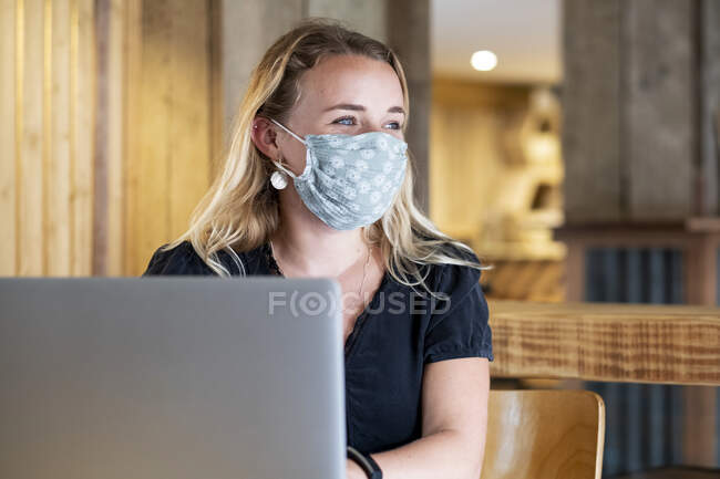 Giovane donna bionda con maschera blu, seduta a tavola, utilizzando il computer portatile. — Foto stock