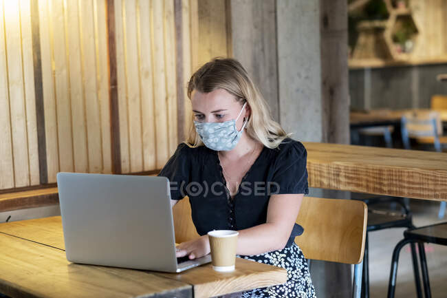 Jovem loira vestindo máscara facial azul, sentada à mesa, usando computador portátil. — Fotografia de Stock