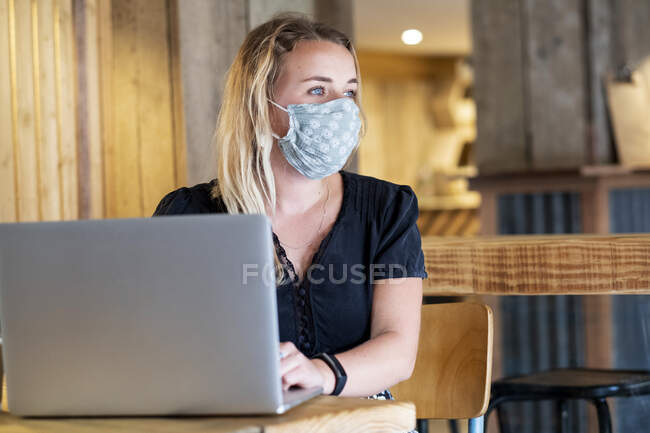 Giovane donna bionda con maschera blu, seduta a tavola, utilizzando il computer portatile. — Foto stock