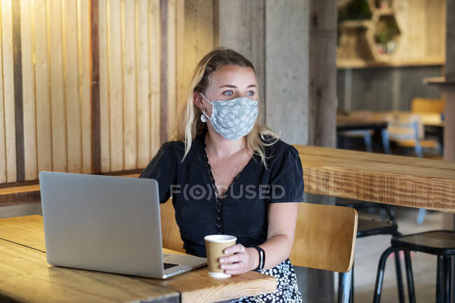 Jeune femme blonde portant un masque bleu, assise à table, à l'aide d'un ordinateur portable. — Photo de stock