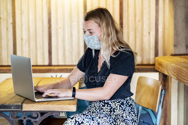 Mujer rubia joven con mascarilla azul, sentada en la mesa, usando computadora portátil. - foto de stock