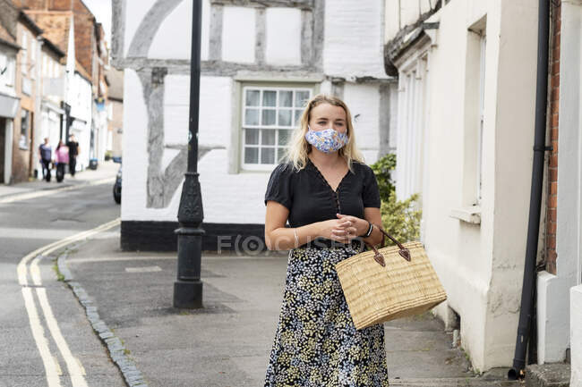 Mujer rubia joven con máscara facial caminando a través de la aldea, llevando bolsa de compras. - foto de stock