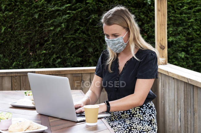 Jovem loira vestindo máscara facial azul, sentada à mesa, usando computador portátil. — Fotografia de Stock