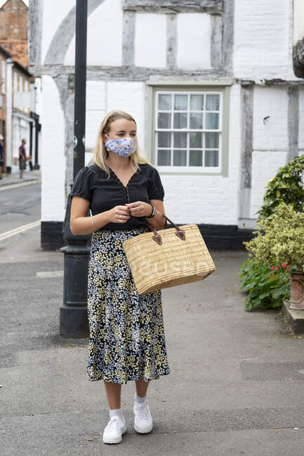 Mujer rubia joven con máscara facial caminando a través de la aldea, llevando bolsa de compras. - foto de stock