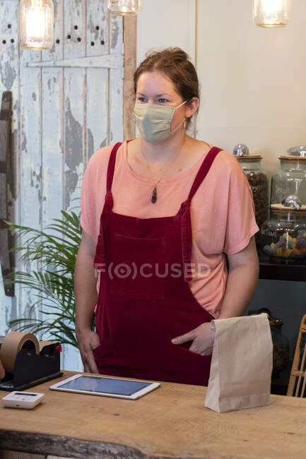 Mujer que usa mascarilla facial y delantal rojo que sirve al cliente en la tienda de productos integrales sin residuos. - foto de stock