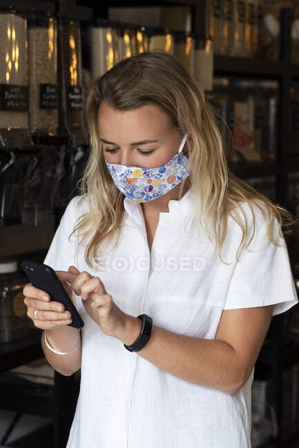 Frau mit Gesichtsmaske kauft mit dem Handy in einem örtlichen Geschäft ein — Stockfoto