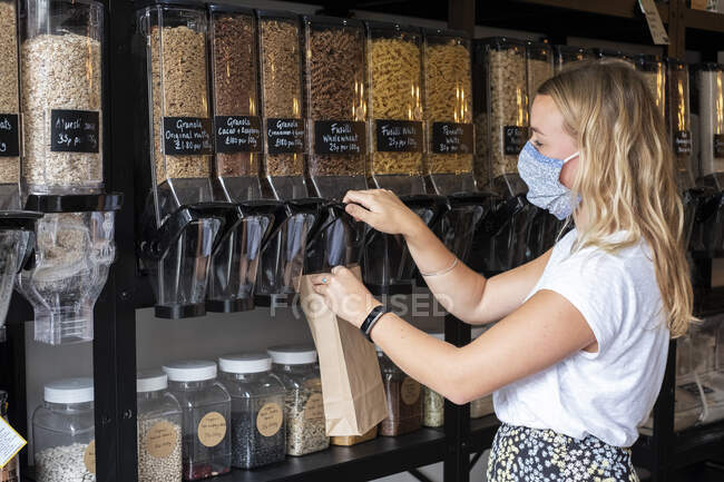 Женщина в маске для лица заполняет бумажный пакет сыпучими ингредиентами — стоковое фото