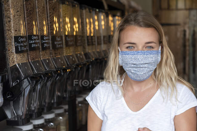 Retrato de una joven rubia con mascarilla facial, parada en una tienda de alimentos integrales sin desperdicios. - foto de stock