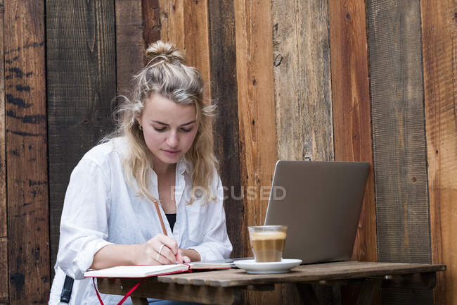 Jeune femme blonde assise seule à une table de café avec un ordinateur portable, écrivant dans un carnet de notes, travaillant à distance. — Photo de stock