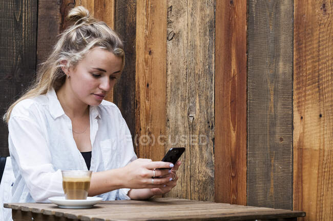 Молода блондинка сидить сама в кафе, використовуючи мобільний телефон, працює дистанційно . — стокове фото